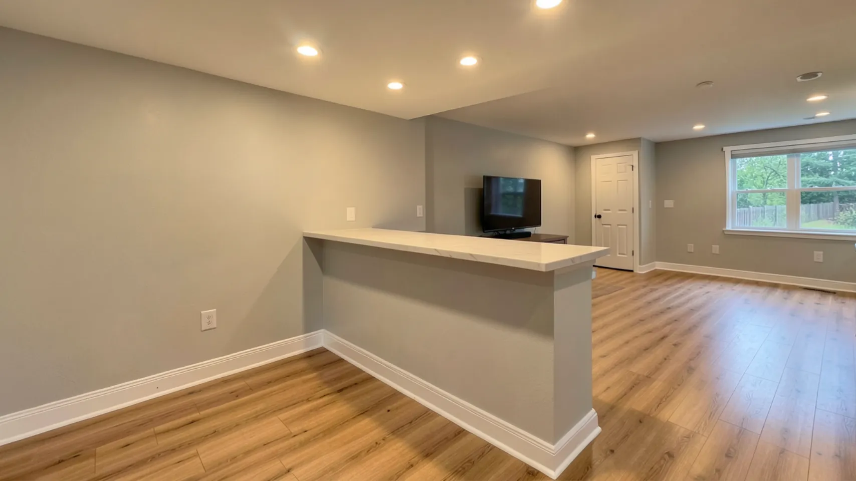 Modern basement remodel in Bloomington featuring a light-colored countertop extending over a partition, with a TV mounted on the wall and natural light from a window, emphasizing functional design and open space.