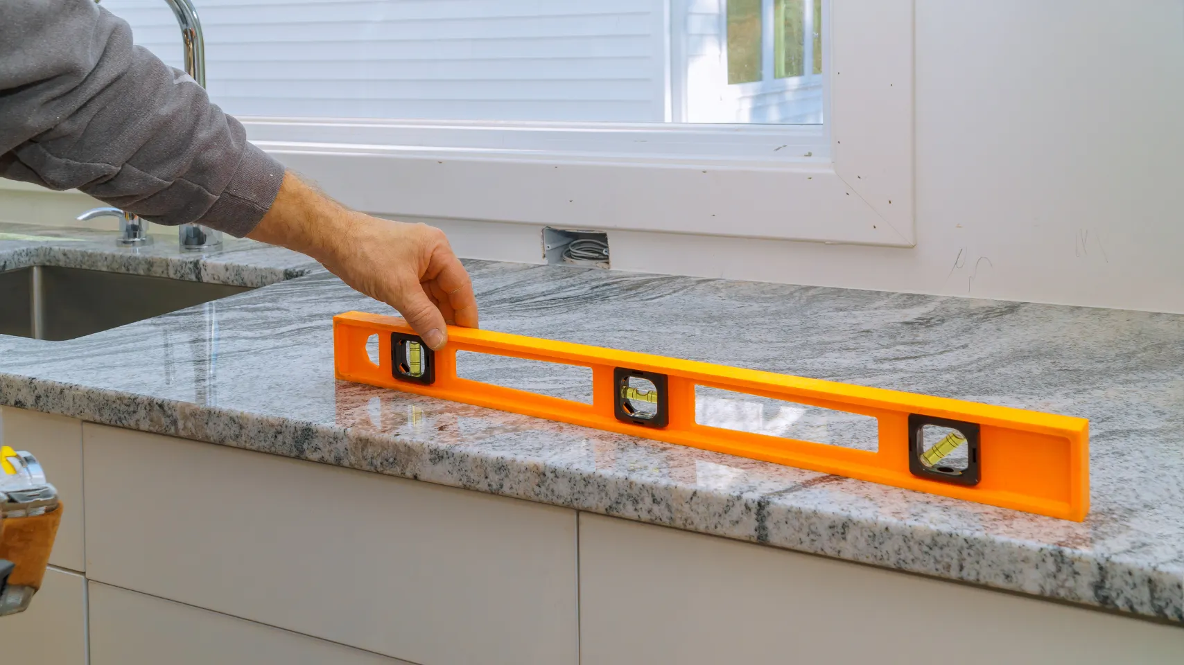 Hand holding an orange level tool on a granite countertop during kitchen remodel, emphasizing accuracy for installation sequencing in Bear Lake projects.
